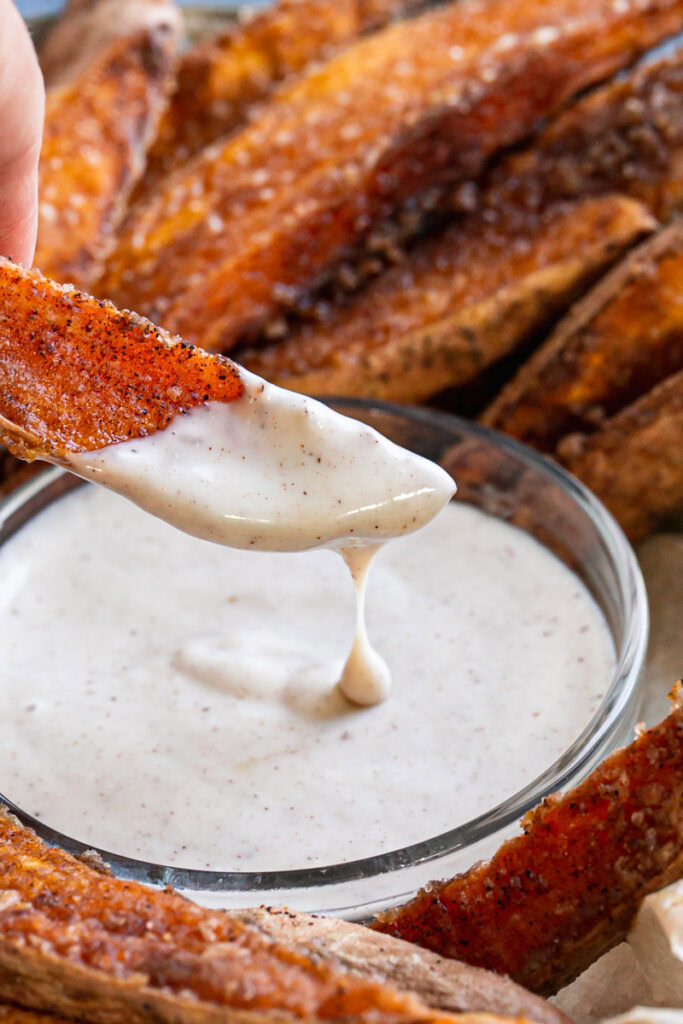 action shot of dipping a crispy cinnamon sugar sweet potato wedge into the coconut sugar sour cream dipping sauce with some dripping off of the wedge into the bowl