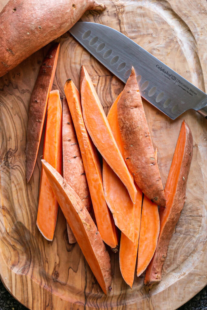a sweet potato sliced into wedges on a wooden cutting board with a whole sweet potato above and the knife resting on the cutting board