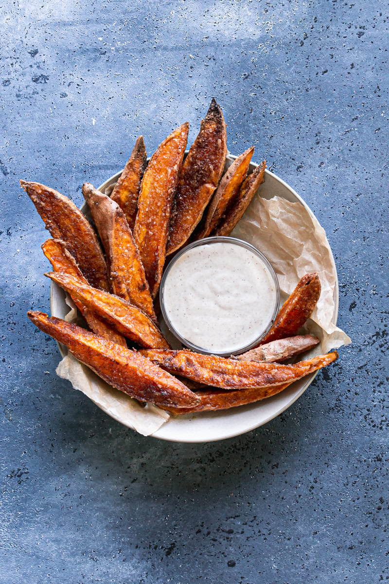 crispy cinnamon sugar sweet potato wedges in a parchment lined bowl surrounding a bowl of coconut sugar sour cream dipping sauce
