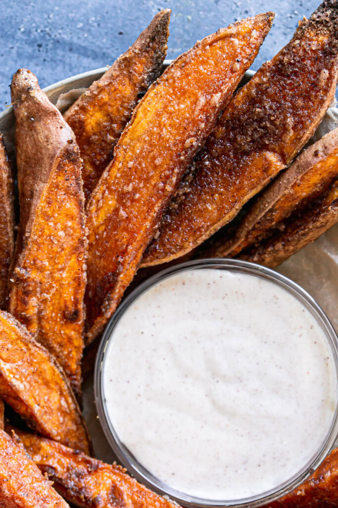 close up shot of crispy cinnamon sugar sweet potato wedges in a parchment lined bowl surrounding a bowl of coconut sugar sour cream dipping sauce