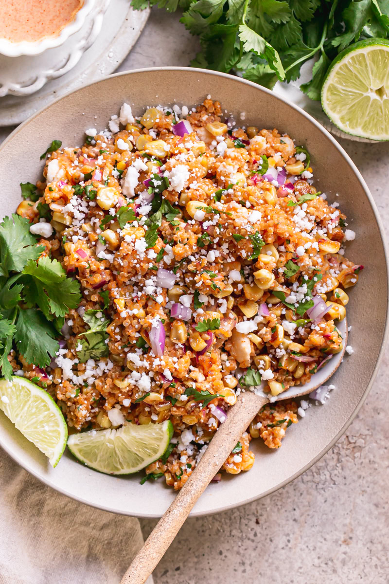 a bowl of Dairy-Free Mexican Street Corn Quinoa Salad with a plate of fresh cilantro leaves and a half of lime above it and a bowl of dressing on the other side