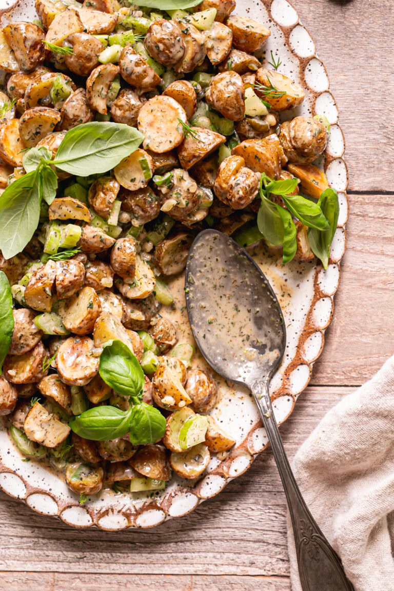 an oval platter with roasted potato salad on it garnished with fresh basil leaves on a wooden table with a spoonful of potato salad missing and the serving spoon on the plate