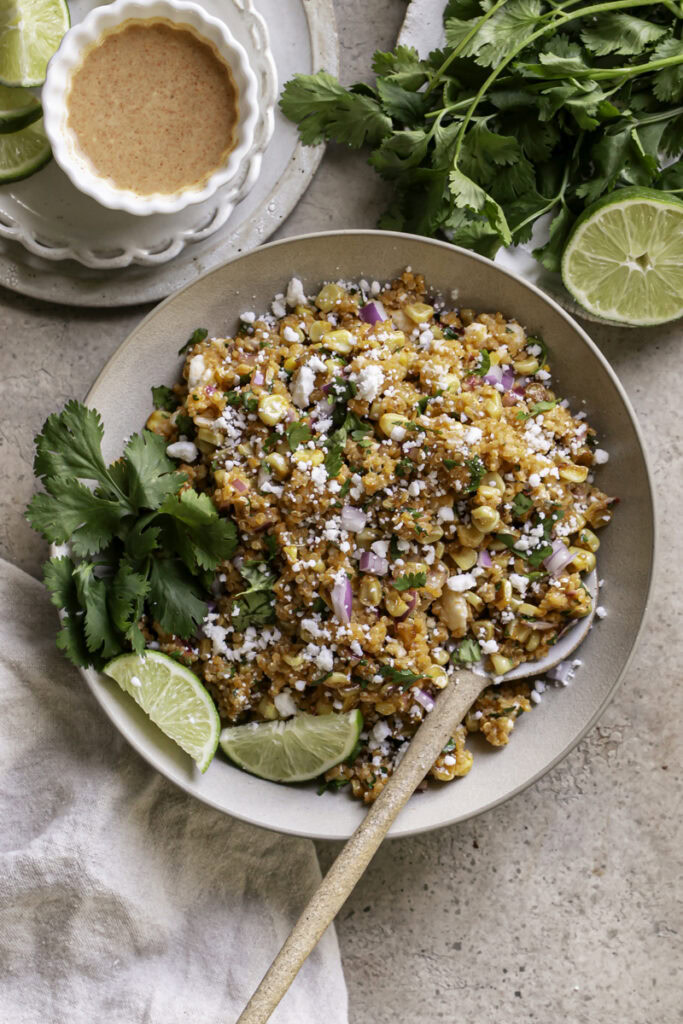 a bowl of Dairy-Free Mexican Street Corn Quinoa Salad with a plate of fresh cilantro leaves and a half of lime above it and a bowl of dressing on the other side.