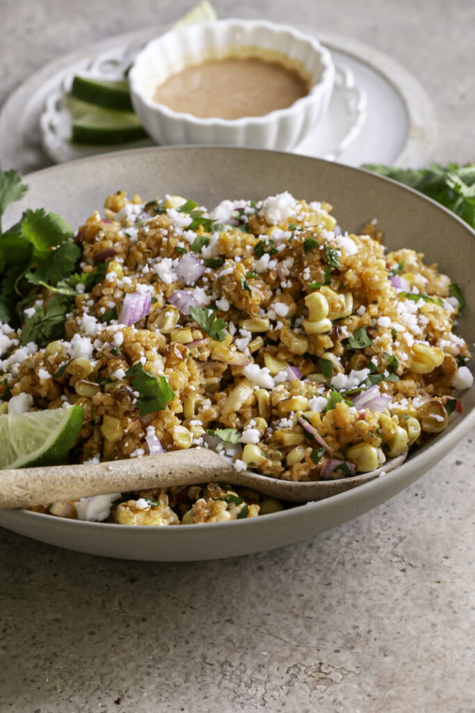 3/4 view of a a bowl of Dairy-Free Mexican Street Corn Quinoa Salad with a spoon in the bowl and a bowl of dressing behind it.