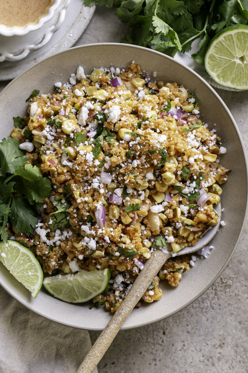 a bowl of Dairy-Free Mexican Street Corn Quinoa Salad with a plate of fresh cilantro leaves and a half of lime above it and a bowl of dressing on the other side.