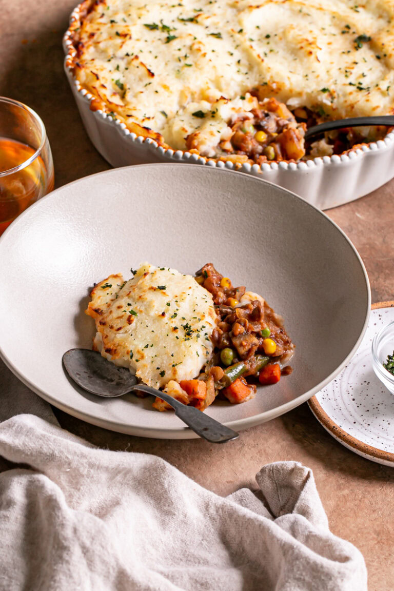 Vegan Gluten-Free Shepherd's Pie served into a bowl with a spoon in it and the pie dish filled with shepherd's pie in the background