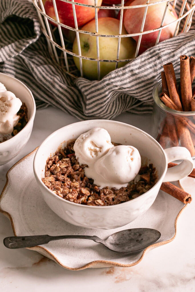 a close up of two mugs of Cinnamon Apple Crisp in a Mug topped with vegan vanilla ice cream, a basket of apples in the background and a jar of cinnamon sticks