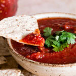 a bowl of restaurant style salsa garnished with fresh cilantro, tortilla chips beside it and one chip dipping into the salsa