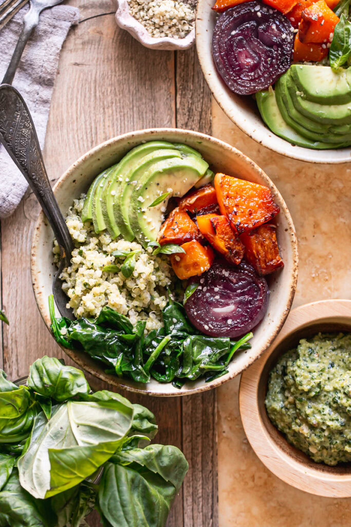 two butternut squash pesto buddha bowls with a spoon in it, a small bowl of pesto below it and a glass of fresh basil