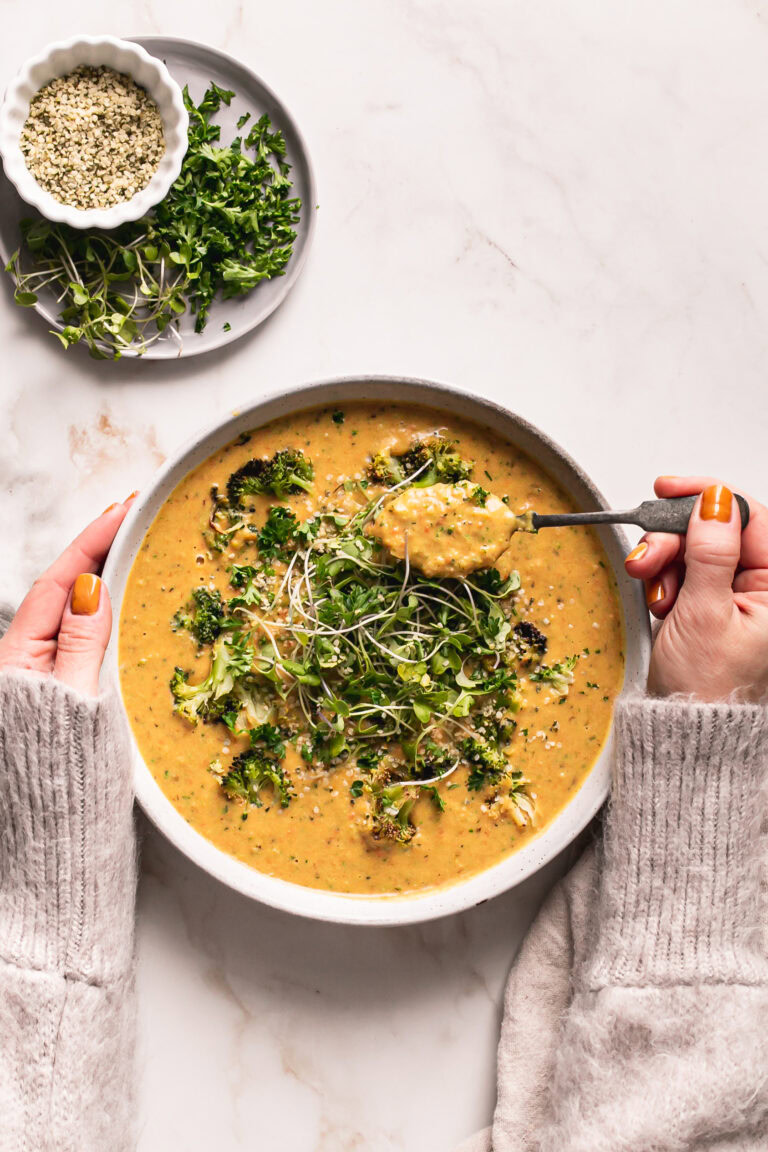 hands holding the bowl of soup scooping a spoonful of soup with a plate of micro greens and help seeds above it