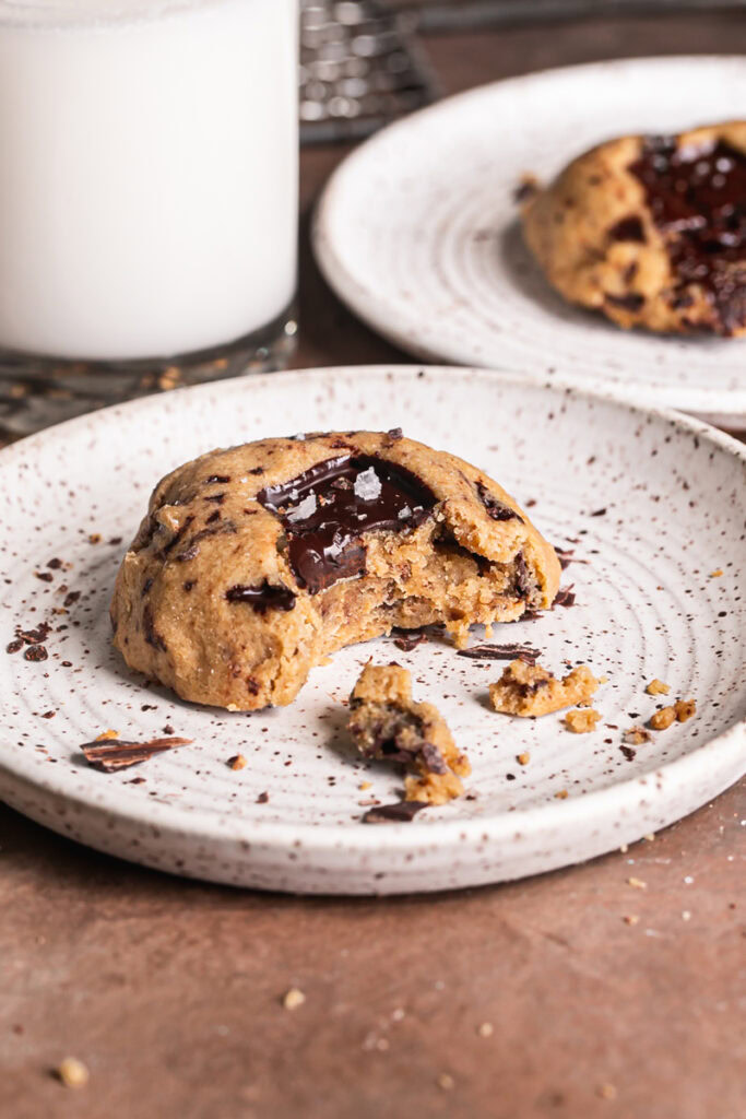 a 3/4 image of a Small batch peanut butter chocolate chunk air fryer cookie on a plate with a bite taken out if it and another cookie on a separate plate in the background with a glass of almond milk