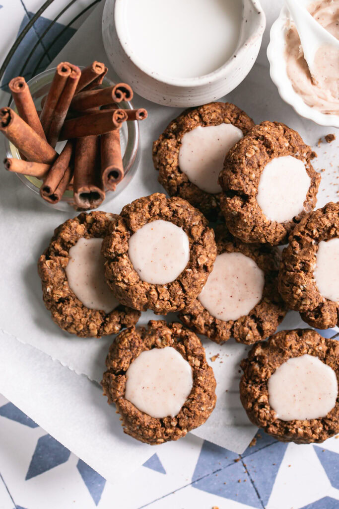 close up over head shot of oatmeal gingerbread thumbprint cookies with white chocolate nog ganache on a parchment-lined round wire cooling rack with a small bowl of white chocolate nog ganache next to it, a jar of milk, and a jar of cinnamon sticks