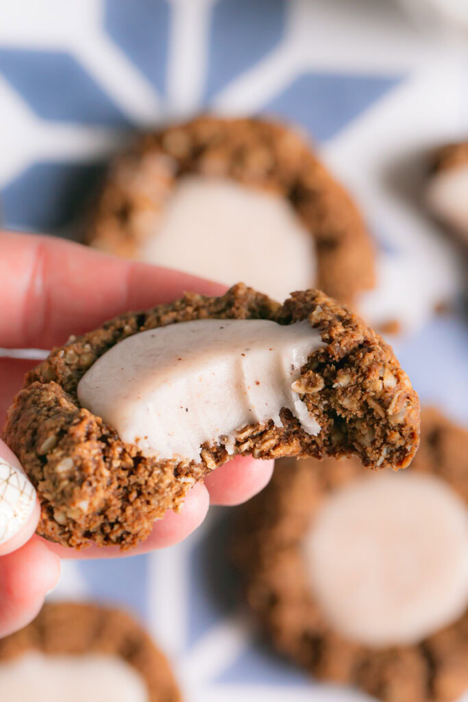 a close up of holding one oatmeal gingerbread thumbprint cookies with white chocolate nog ganache cookie with a bite taken out of it