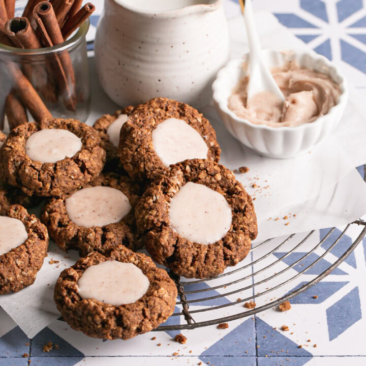 oatmeal gingerbread thumbprint cookies with white chocolate nog ganache on a parchment-lined round wire cooling rack with a small bowl of white chocolate nog ganache next to it, a jar of milk, and a jar of cinnamon sticks