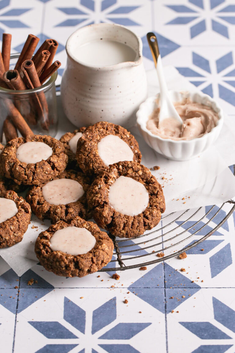 oatmeal gingerbread thumbprint cookies with white chocolate nog ganache on a parchment-lined round wire cooling rack with a small bowl of white chocolate nog ganache next to it, a jar of milk, and a jar of cinnamon sticks