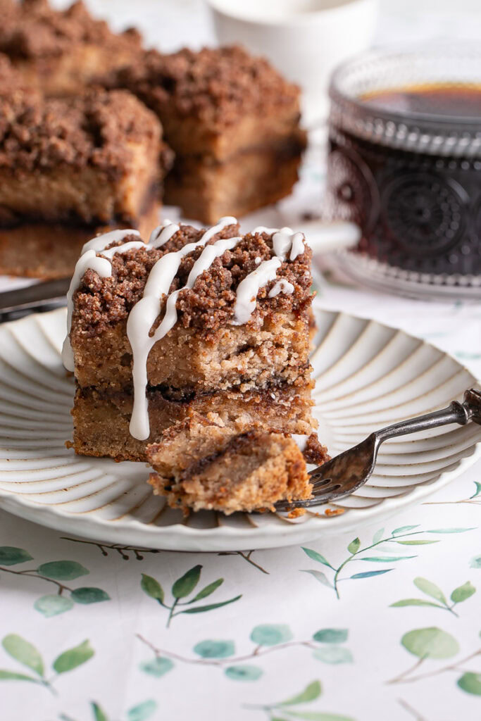 a piece of almond flour coffee cake drizzled with icing, a little piece on a fork with the rest of the coffee cake and a mug of coffee in the background