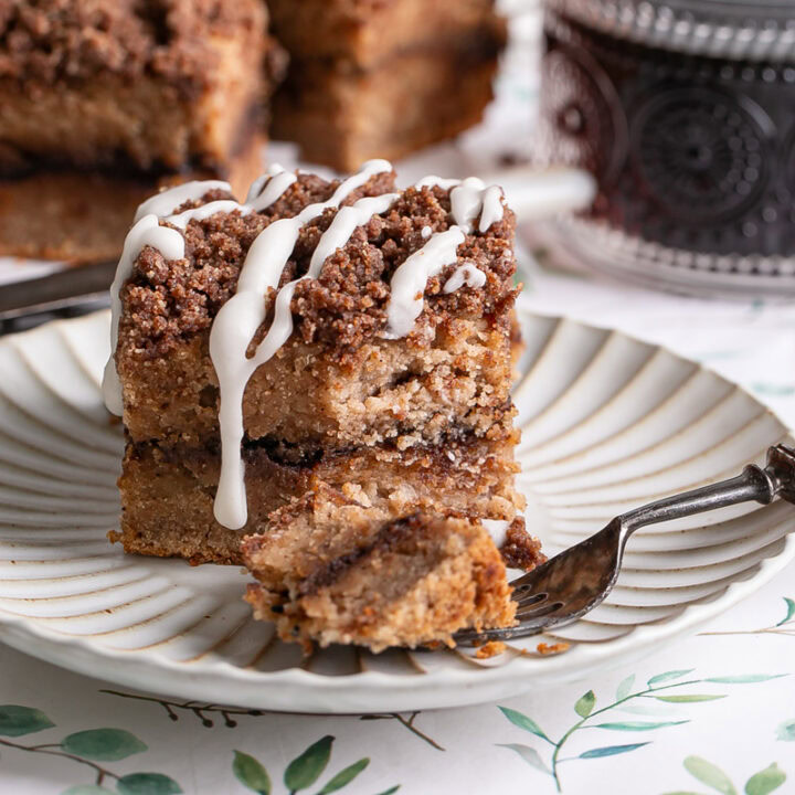 a piece of almond flour coffee cake drizzled with icing, a little piece on a fork with the rest of the coffee cake and a mug of coffee in the background