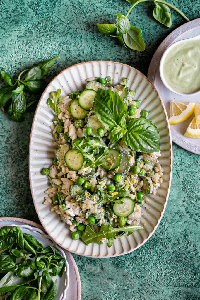 an oval plate full of lemon basil orzo salad garnished with fresh basil, fresh basil leaves laid around the plate, another plate with a bowl of lemon basil tahini dressing and lemon slices, and a plate of fresh basil leaves in the bottom left corner