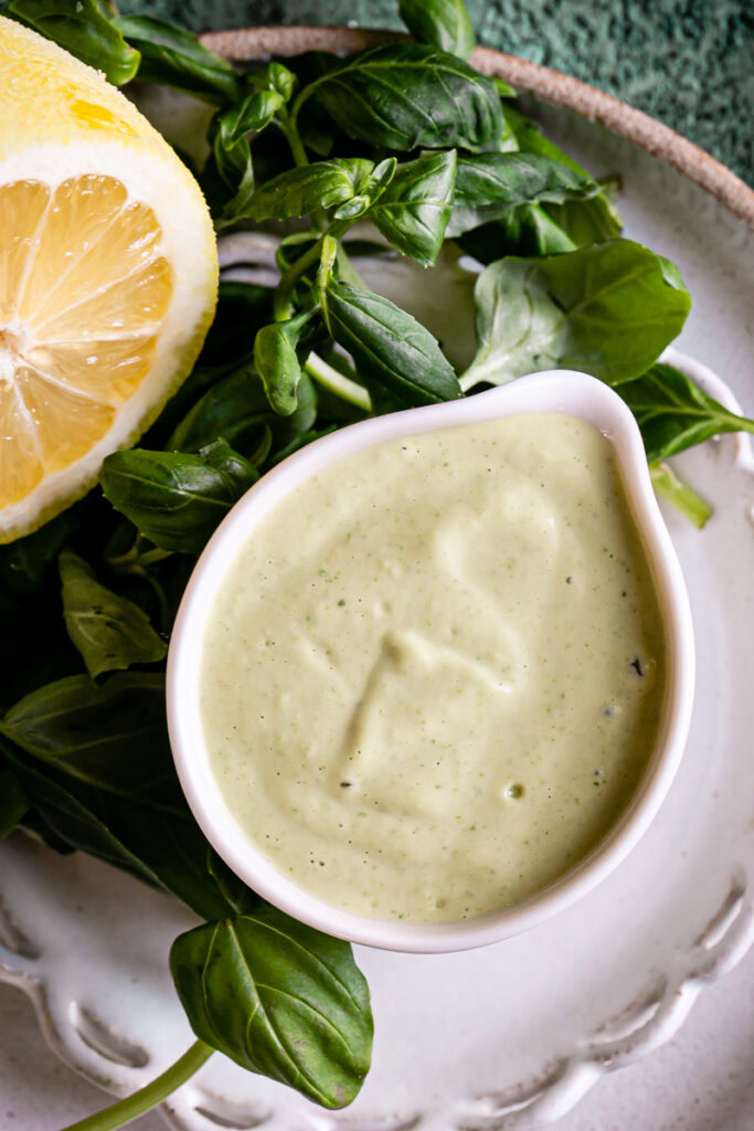 a white measuring cup full of lemon basil tahini dressing with fresh basil leaves and a sliced open lemon on the plate next to the dressing