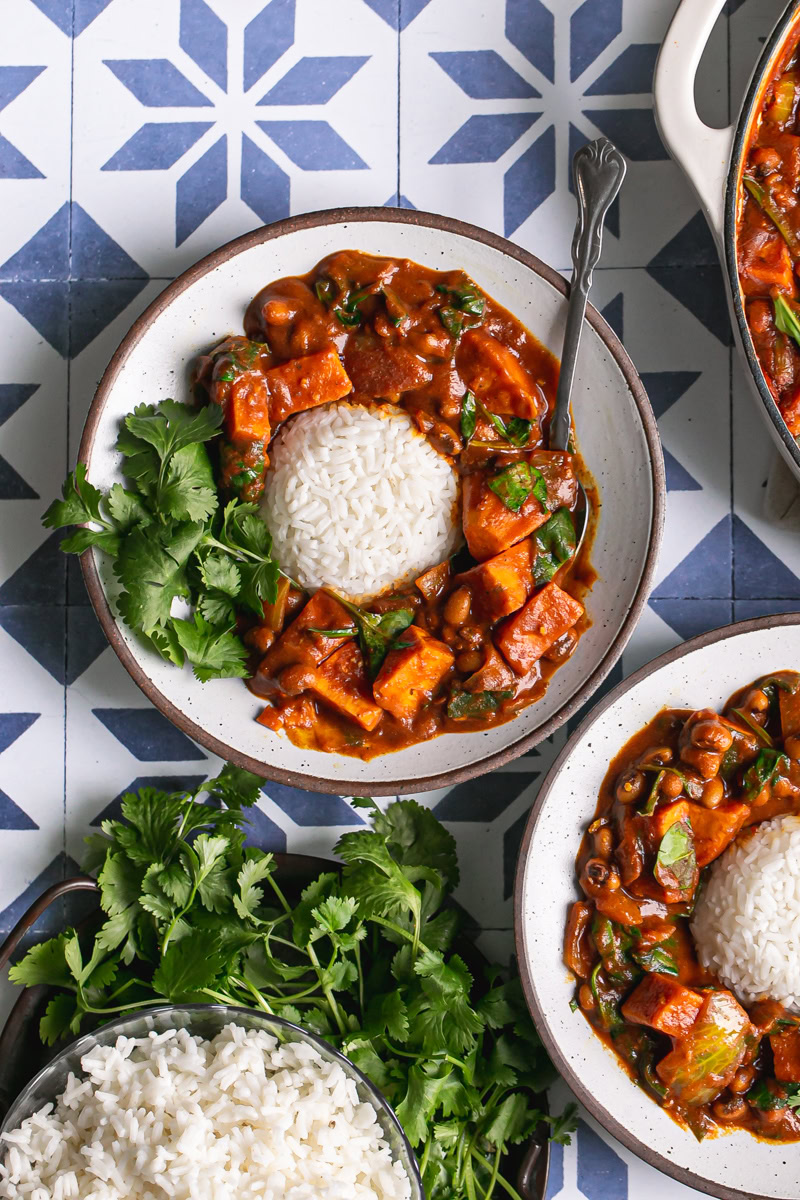 two bowls of close up of a wooden serving spoon scooping up some sweet potato black eyed pea curry with the white rice in the middle and the curry around the rice, one bowl with a spoon in it, and a plate of fresh cilantro
