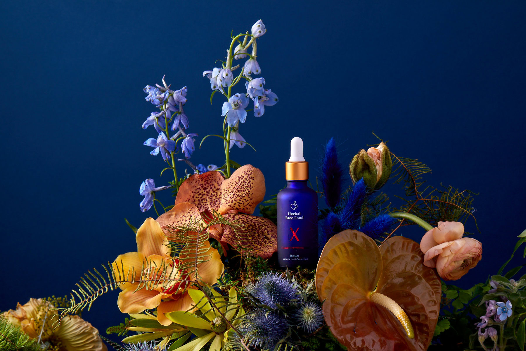 a bottle of herbal face food the cure x serum on a blue backdrop with exotic plants and flowers