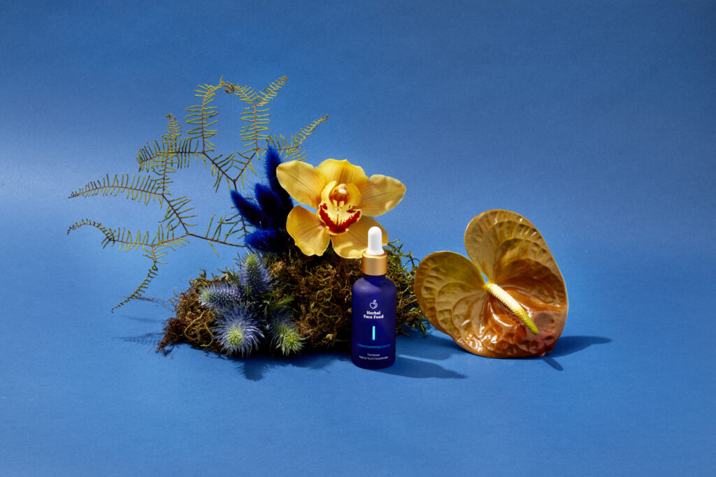 a bottle of herbal face food serum 1 on a blue backdrop with exotic plants and flowers
