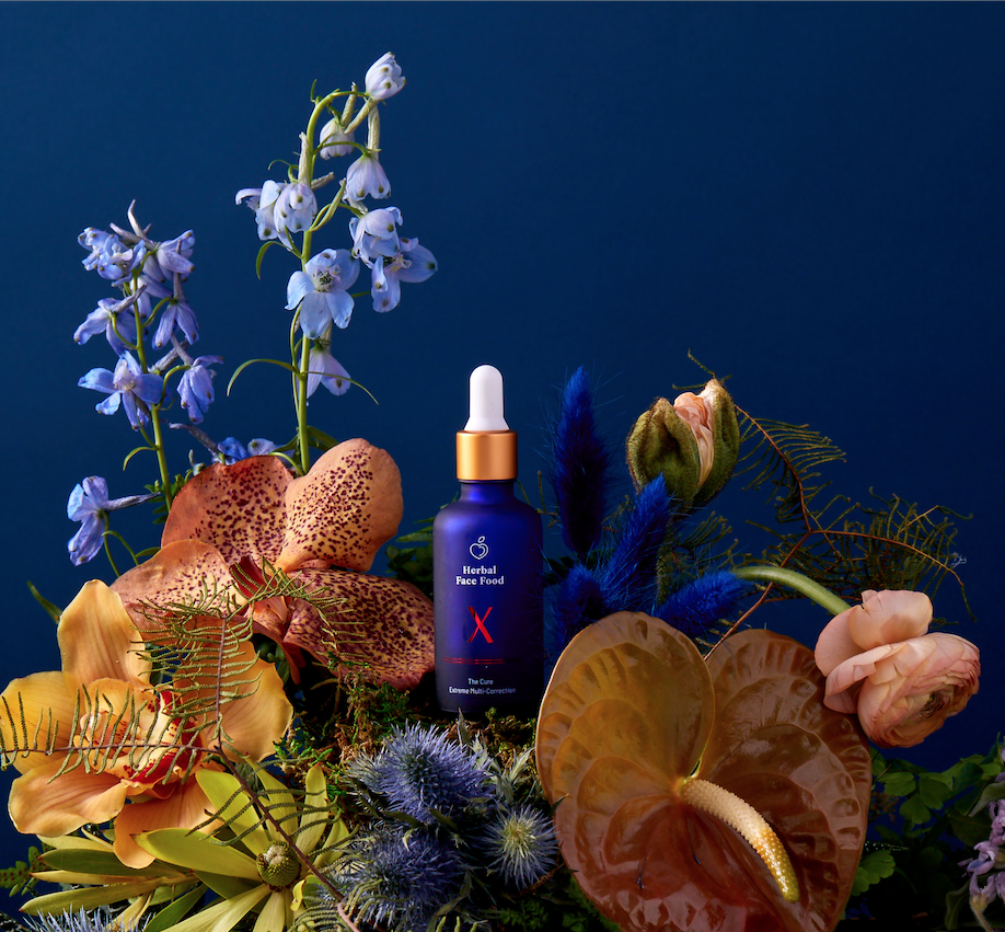 a bottle of herbal face food the cure x serum on a blue backdrop with exotic plants and flowers