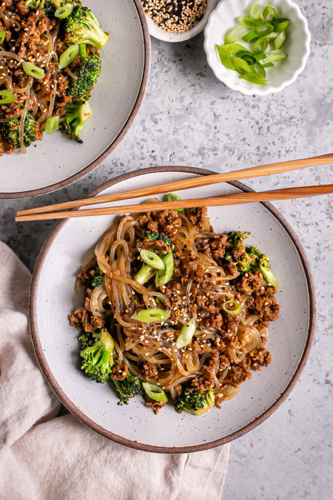 overhead image of two bowls of vegan beef and broccoli teriyaki noodles garnished with green onions and sesame seeds with chopsticks resting on one of the bowls