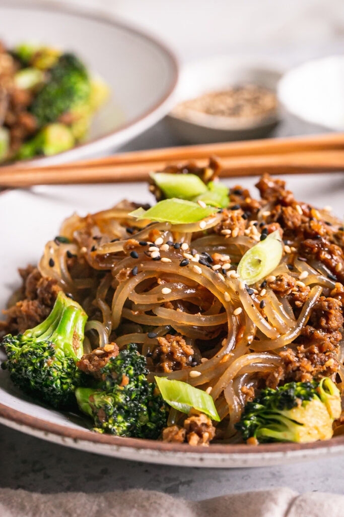 a very close up image of the vegan beef and broccoli teriyaki noodles in a bowl with chopsticks resting on the bowl