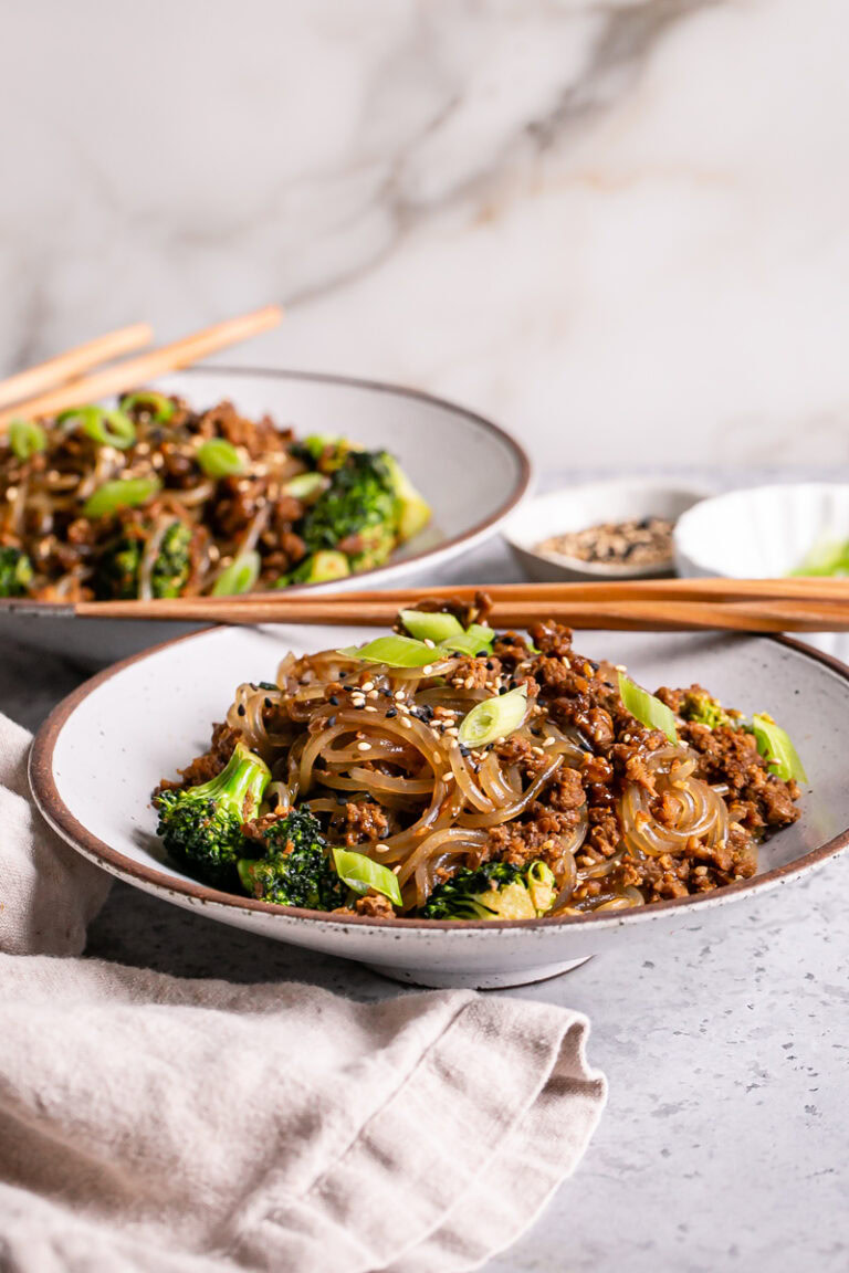 two bowls of vegan beef and broccoli teriyaki noodles with chopsticks laying on th bowls and a linen napkin in front of the first bowl