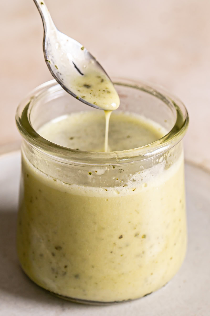 close up of a jar of lemon basil vinaigrette dressing with a spoon drizzling some dressing back into the jar