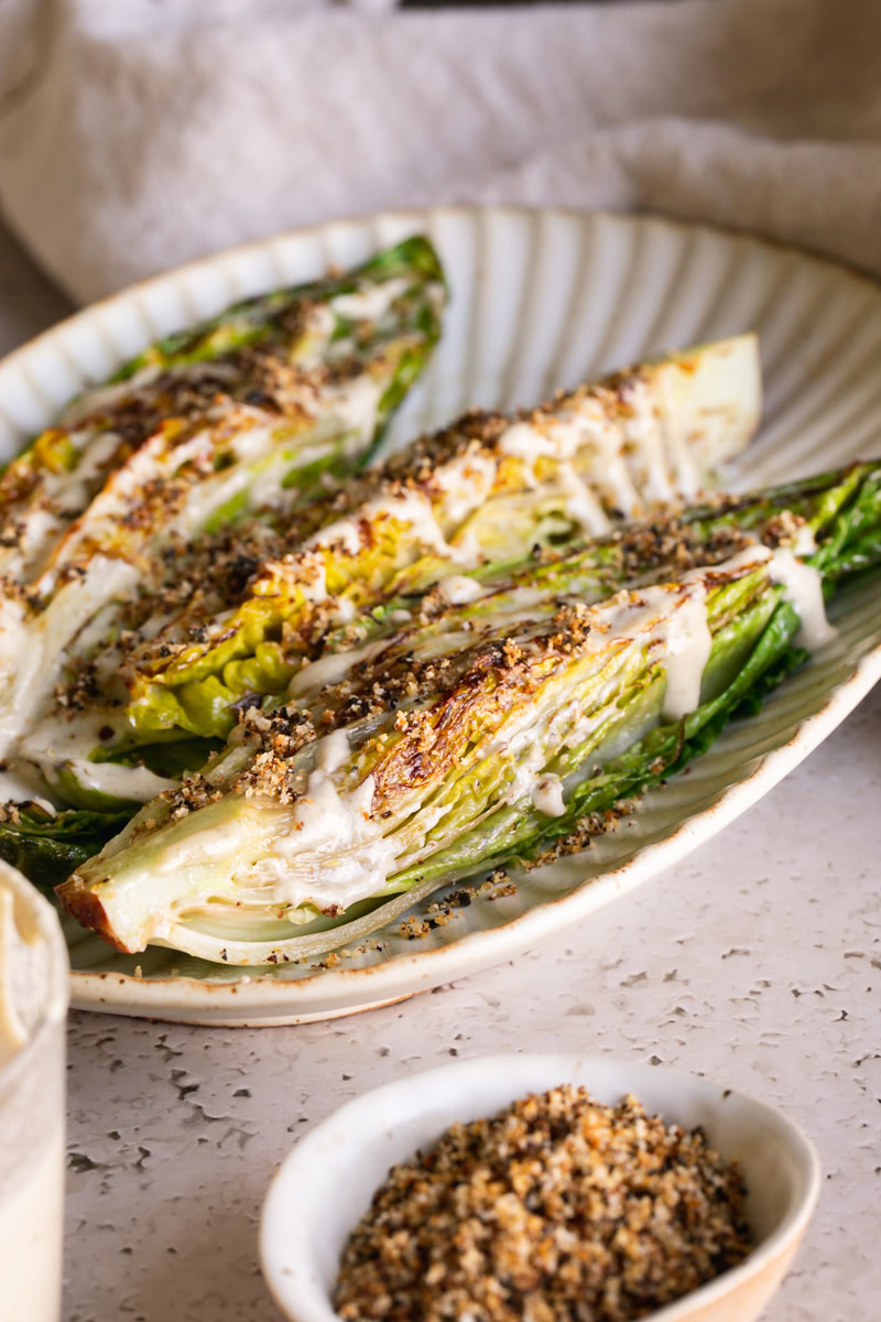 3/4 shot of three halves of romaine lettuce heads on an oval plate drizzled with caesar dressing and sprinkled with gluten-free toasted breadcrumbs.