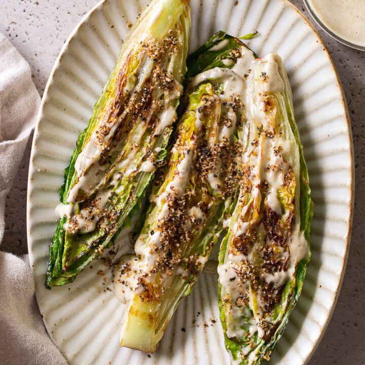 three halves of romaine lettuce heads on an oval plate drizzled with caesar dressing and sprinkled with gluten-free toasted breadcrumbs. A bowl of bread crumbs and caesar dressing above the oval plate.