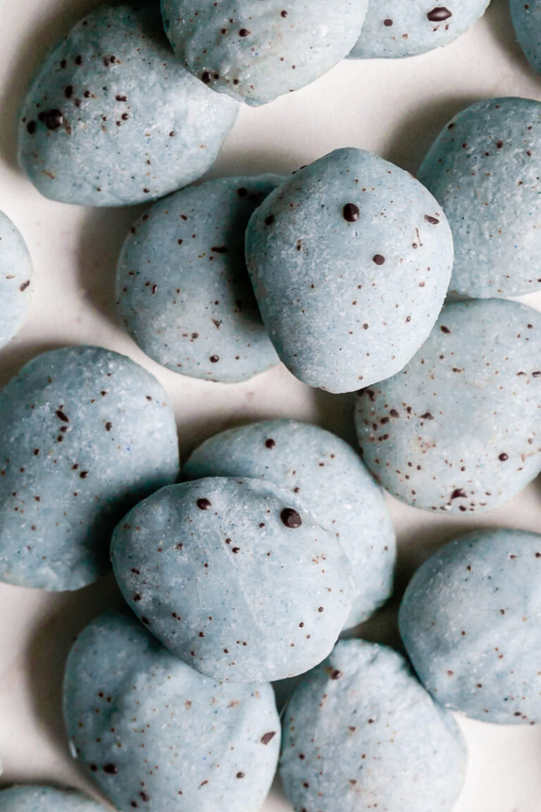 a close up of a pile of white chocolate covered almonds colored robin's egg blue using blue spirulina and speckled with dark chocolate.