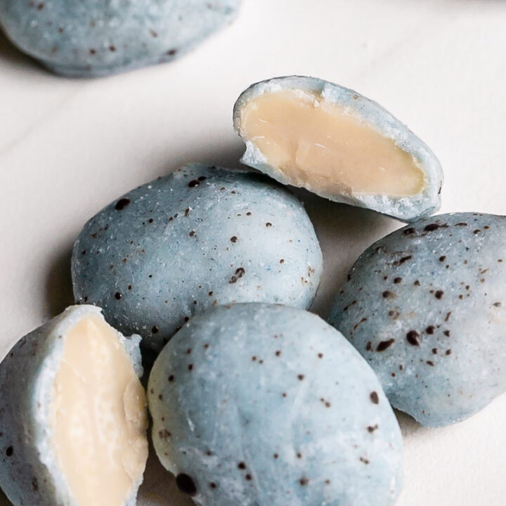 a close up of a pile of white chocolate covered almonds colored robin's egg blue using blue spirulina and speckled with dark chocolate with two almonds cut in half to reveal the almond inside