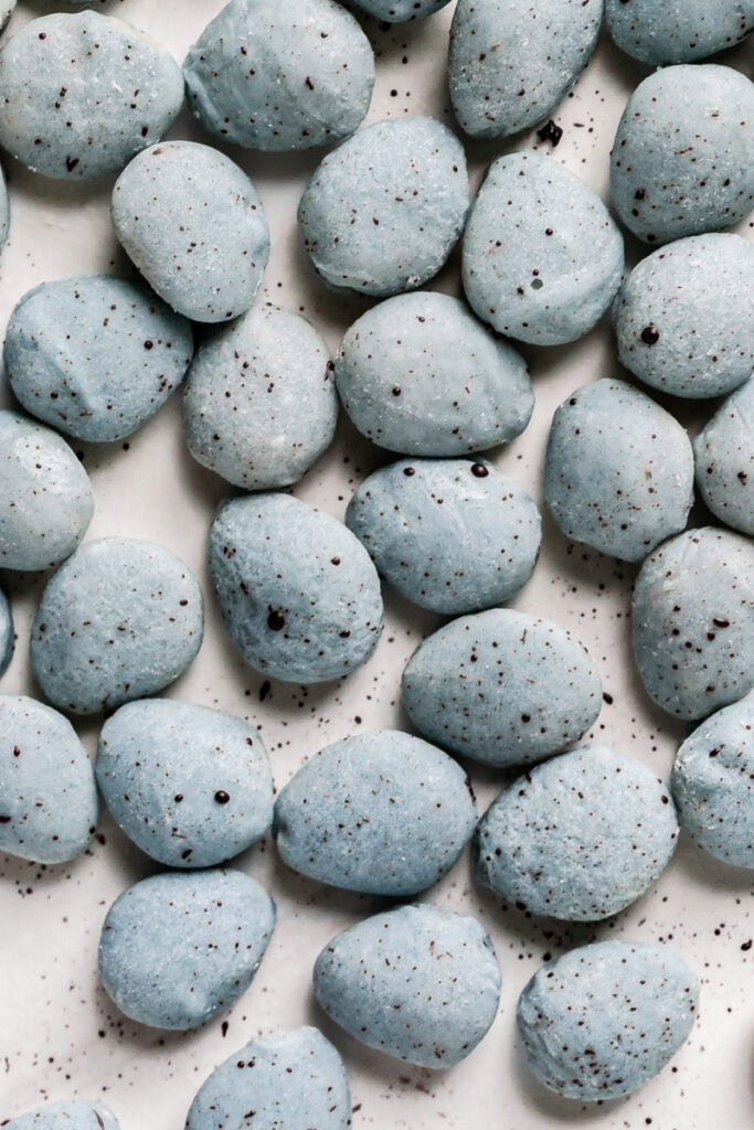 a pile of white chocolate covered almonds colored robin's egg blue using blue spirulina and speckled with dark chocolate