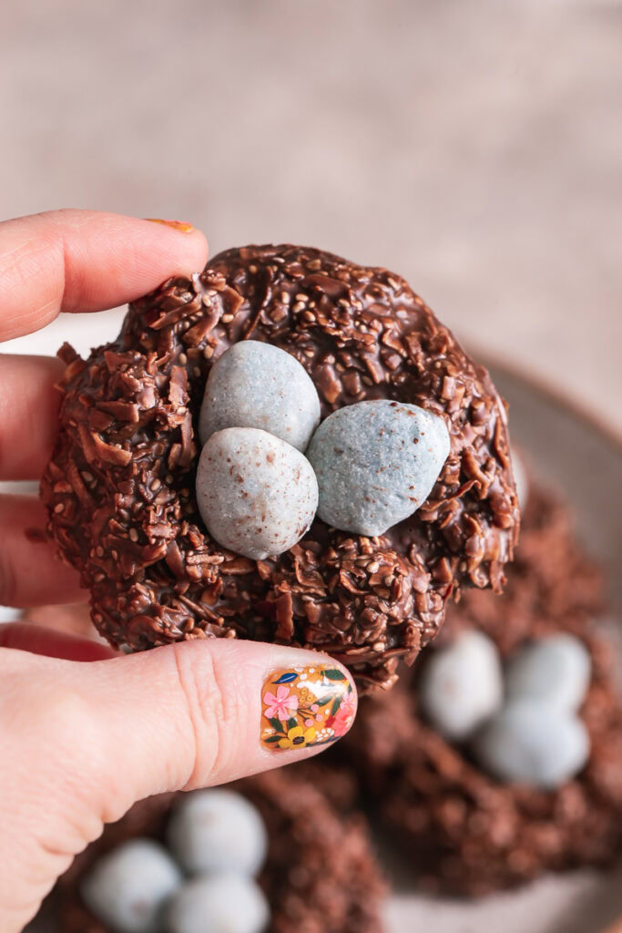 a hand holding one of the 3/4 shot of a Toasted Coconut Chocolate Bird's Egg Nest Cookie filled with three robin's egg white chocolate covered almonds in it and other cookies in the background