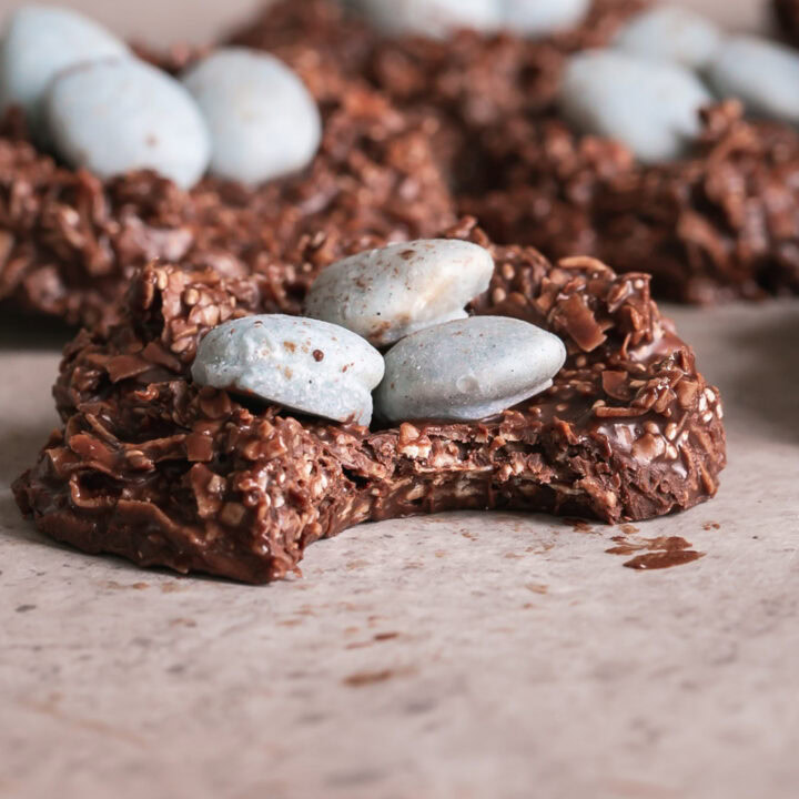 3/4 shot of a Toasted Coconut Chocolate Bird's Egg Nest Cookie filled with three robin's egg white chocolate covered almonds in it and a bite taken out of the cookie with other cookies in the background