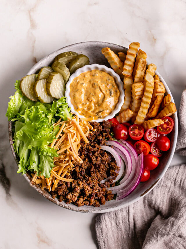 a bowl filled with lettuce, plant-based ground beef, French fries, cherry tomatoes, sliced red onion, vegan shredded cheddar cheese, dill pickle chips, and a bowl of homemade dressing to make this Vegan Cheeseburger Salad