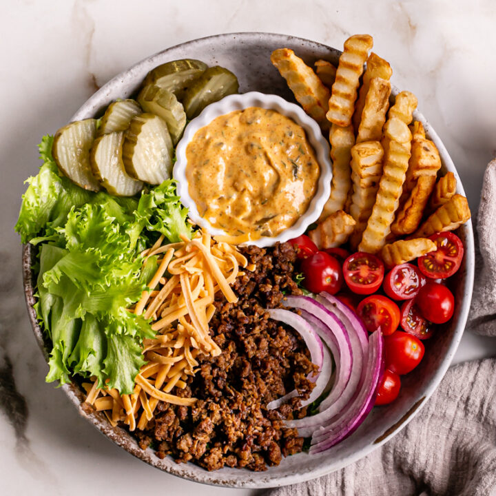 a bowl filled with lettuce, plant-based ground beef, French fries, cherry tomatoes, sliced red onion, vegan shredded cheddar cheese, dill pickle chips, and a bowl of homemade dressing to make this Vegan Cheeseburger Salad
