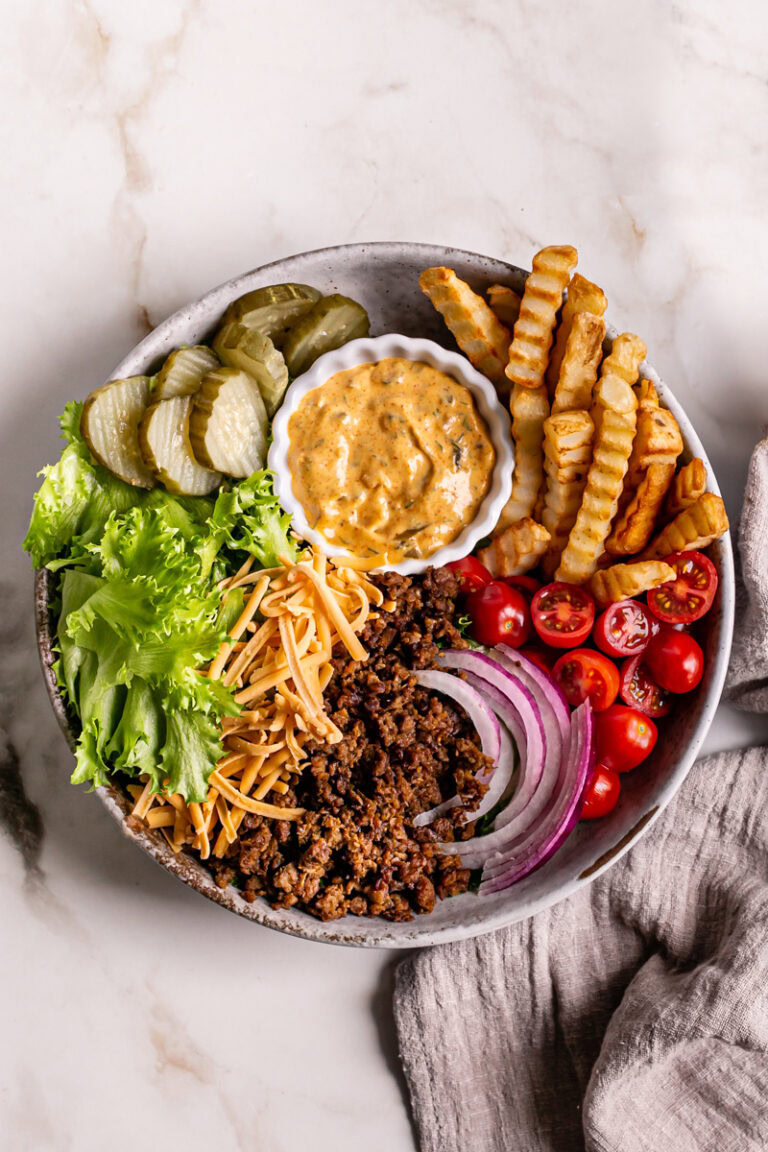 a bowl filled with lettuce, plant-based ground beef, French fries, cherry tomatoes, sliced red onion, vegan shredded cheddar cheese, dill pickle chips, and a bowl of homemade dressing to make this Vegan Cheeseburger Salad