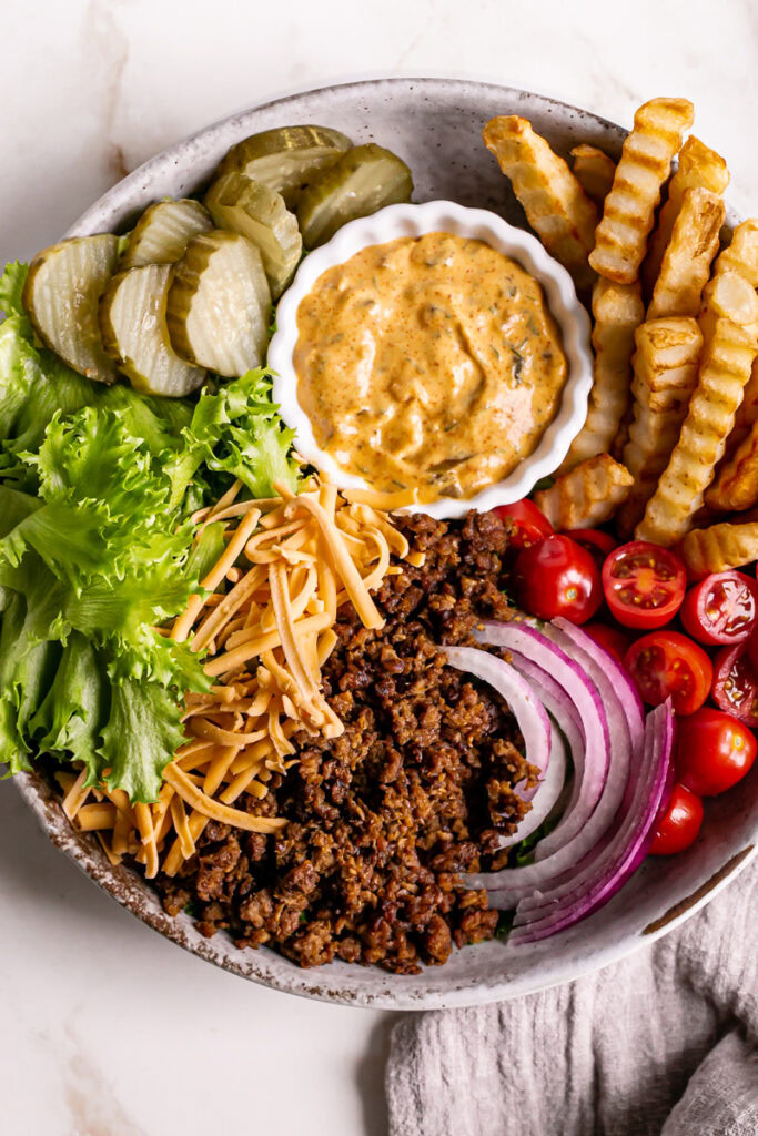 a close up of a bowl filled with lettuce, plant-based ground beef, French fries, cherry tomatoes, sliced red onion, vegan shredded cheddar cheese, dill pickle chips, and a bowl of homemade dressing to make this Vegan Cheeseburger Salad