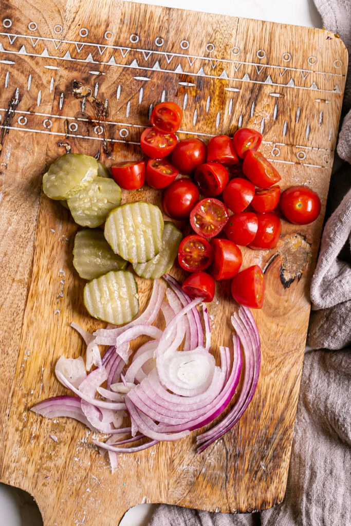 sliced red onion, cherry tomatoes cut in half, and dill pickle chips on a wooden cutting board