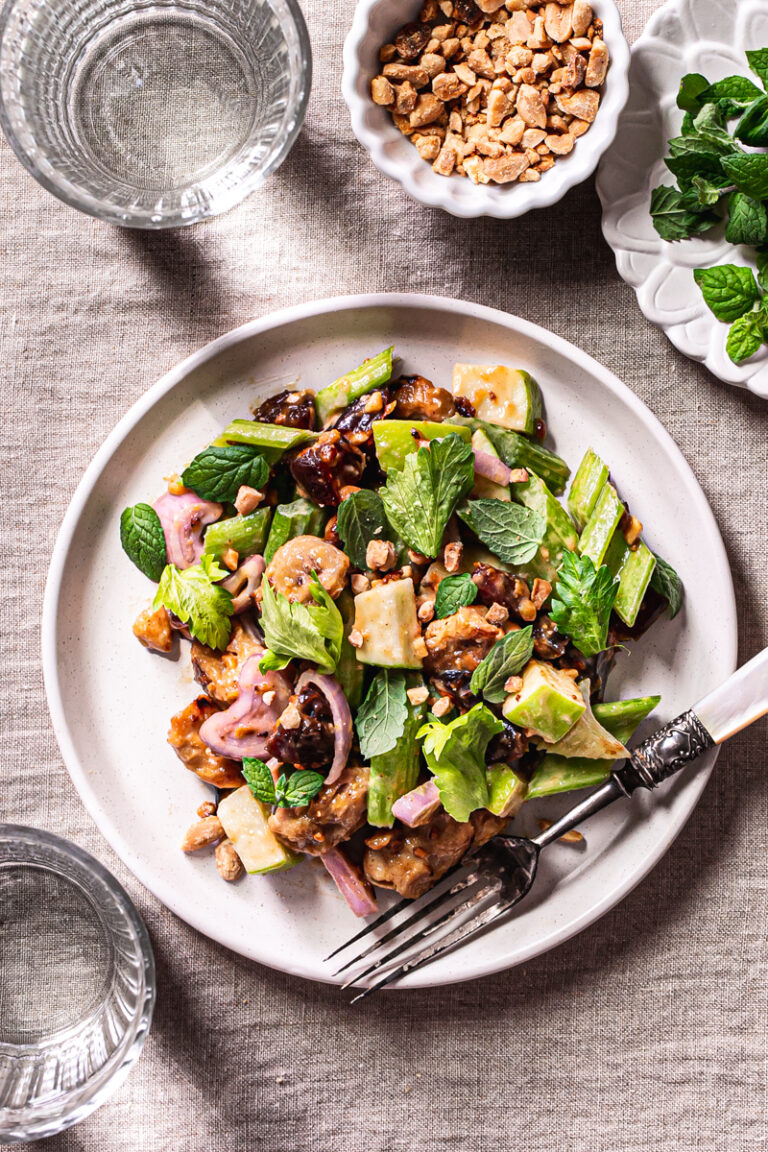 a plate of Ants On A Log Celery date salad with plant-based Chicken, a fork on the plate, a bowl or crushed roasted peanuts, a small plate of fresh mint, and two glasses of water on a linen tablecloth