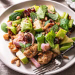 3/4 shot of a plate of Ants On A Log Celery date salad with plant-based Chicken, a fork on the plate, a bowl or crushed roasted peanuts, a small plate of fresh mint, and two glasses of water on a linen tablecloth
