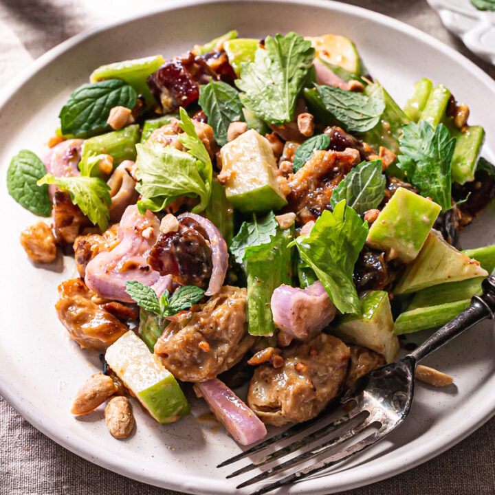 3/4 shot of a plate of Ants On A Log Celery date salad with plant-based Chicken, a fork on the plate, a bowl or crushed roasted peanuts, a small plate of fresh mint, and two glasses of water on a linen tablecloth