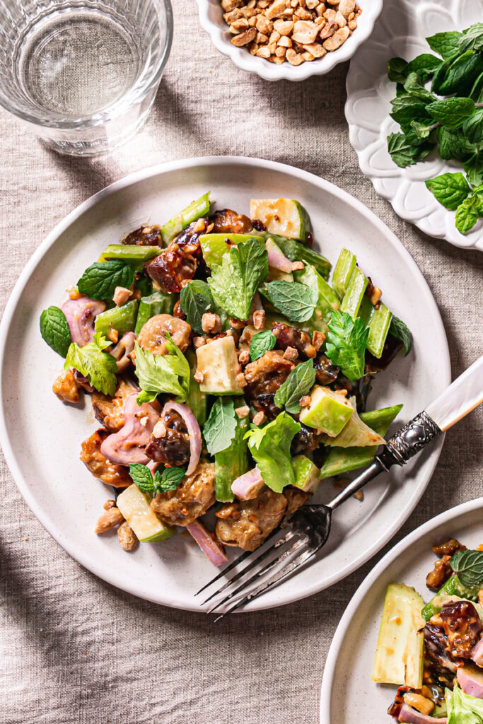 a plate of Ants On A Log Celery date salad with plant-based Chicken, a fork on the plate, a bowl or crushed roasted peanuts, a small plate of fresh mint, and two glasses of water on a linen tablecloth