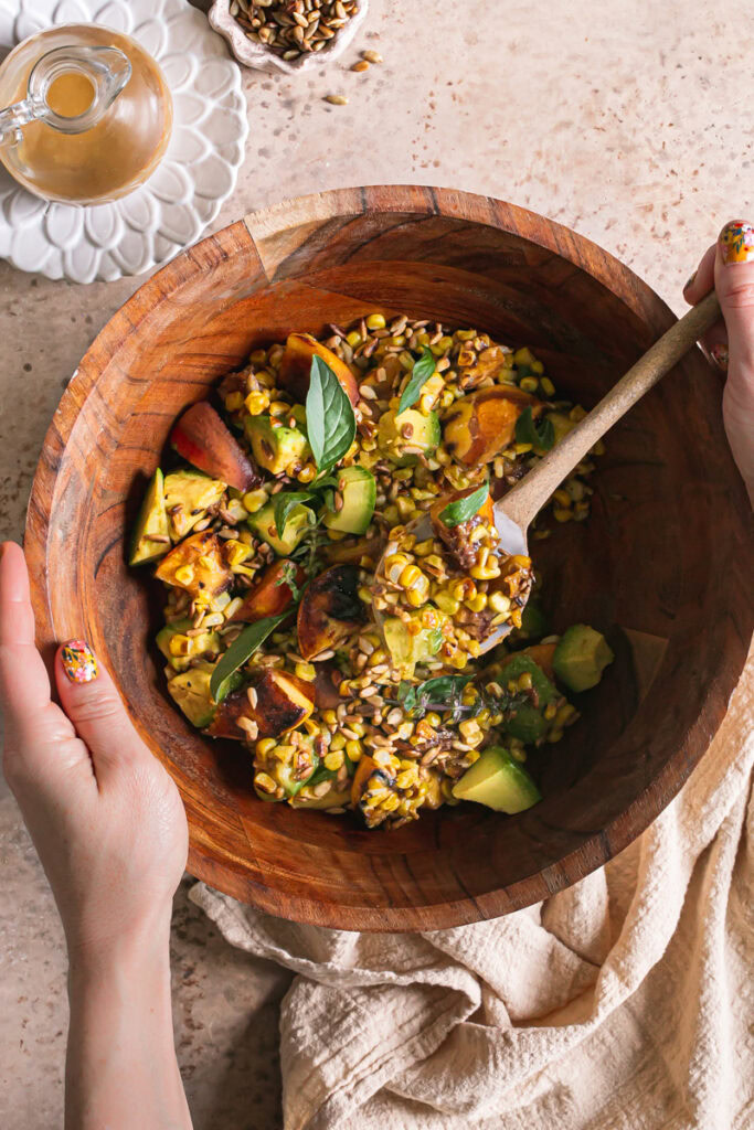 hands holding a wooden bowl with Grilled Corn Peach Avocado Salad in it and scooping some of the salad out of the bowl. A plate of Orange Ginger Miso Dressing in a glass dressing bottle on the side and a small bowl of toasted sunflower seeds beside it