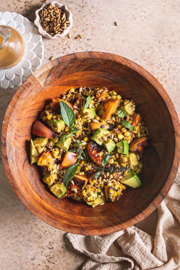 Grilled Corn Peach Avocado Salad with Orange Ginger Miso Dressing in a wooden bowl, a linen napkin under the bowl, a small bowl of toasted sunflower seeds above the wooden bowl and a plate with a glass bottle of orange ginger miso dressing