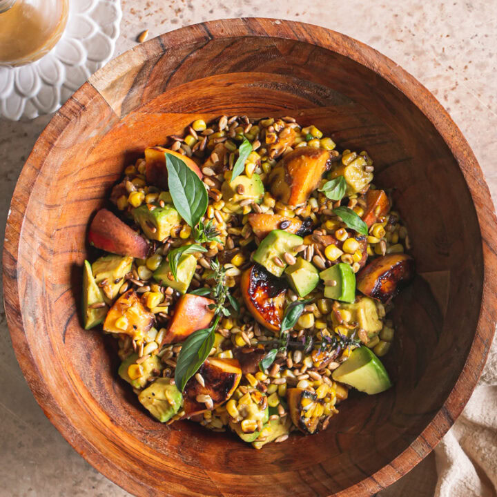 Grilled Corn Peach Avocado Salad with Orange Ginger Miso Dressing in a wooden bowl, a linen napkin under the bowl, a small bowl of toasted sunflower seeds above the wooden bowl and a plate with a glass bottle of orange ginger miso dressing